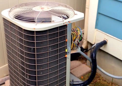 An outdoor air conditioner condenser beside a house, its wiring panel open for service.